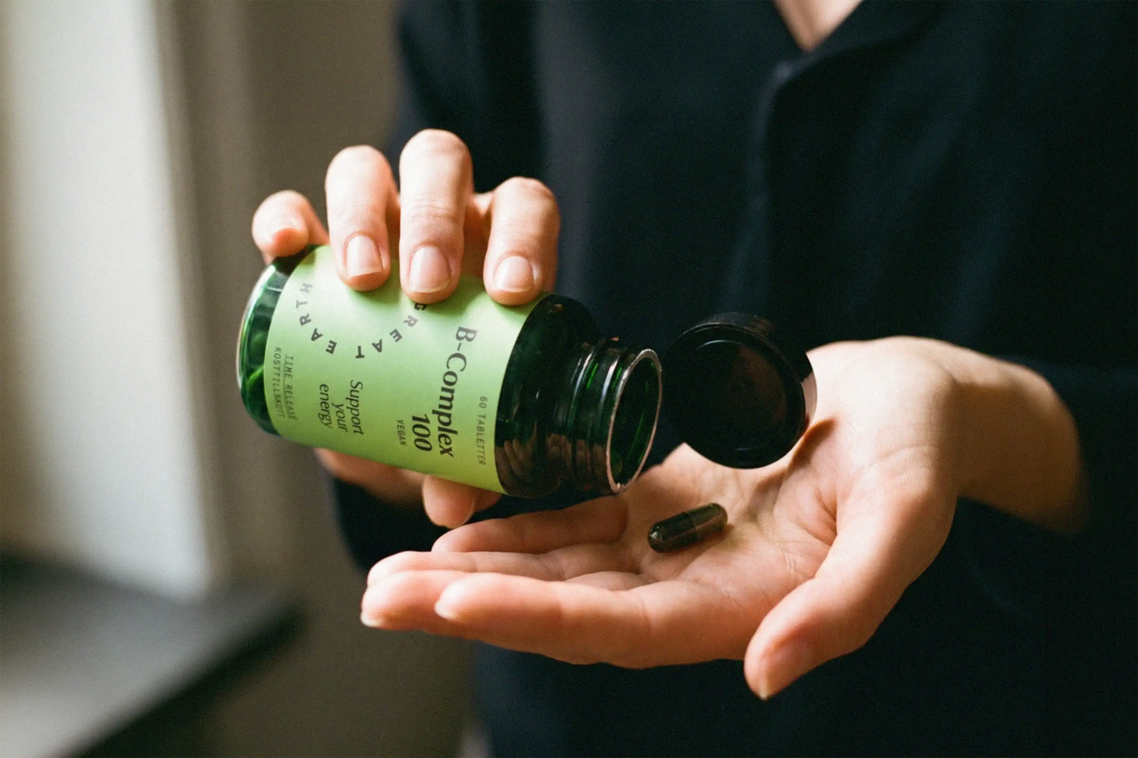 Hand håller upp flaska med B-Complex 100 B-vitamintillskott från Great Earth – veganskt kosttillskott med tidsreglerad frisättning