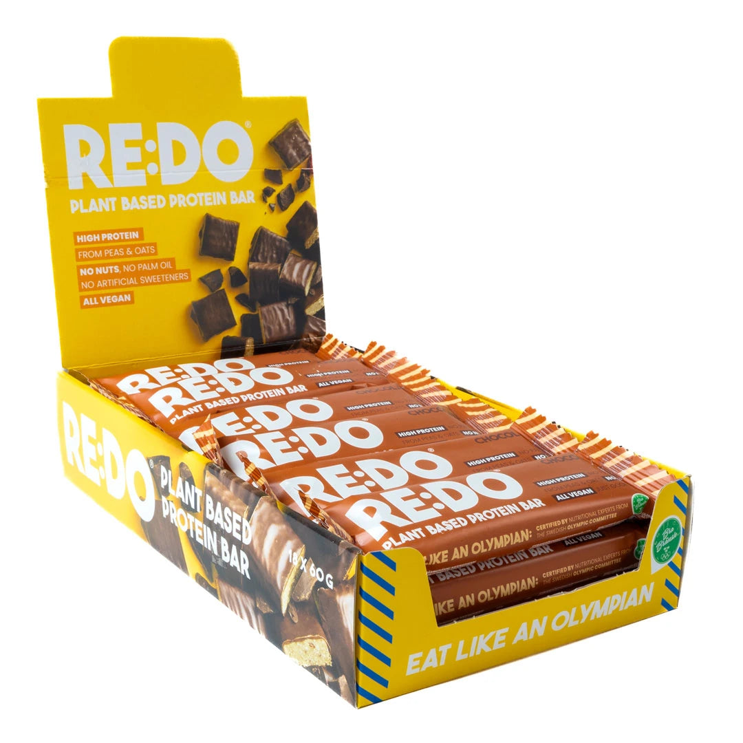 18 x REDO Bar 60 g Chocolate Chock i en öppen gul displaylåda fylld med bruna proteinbars.