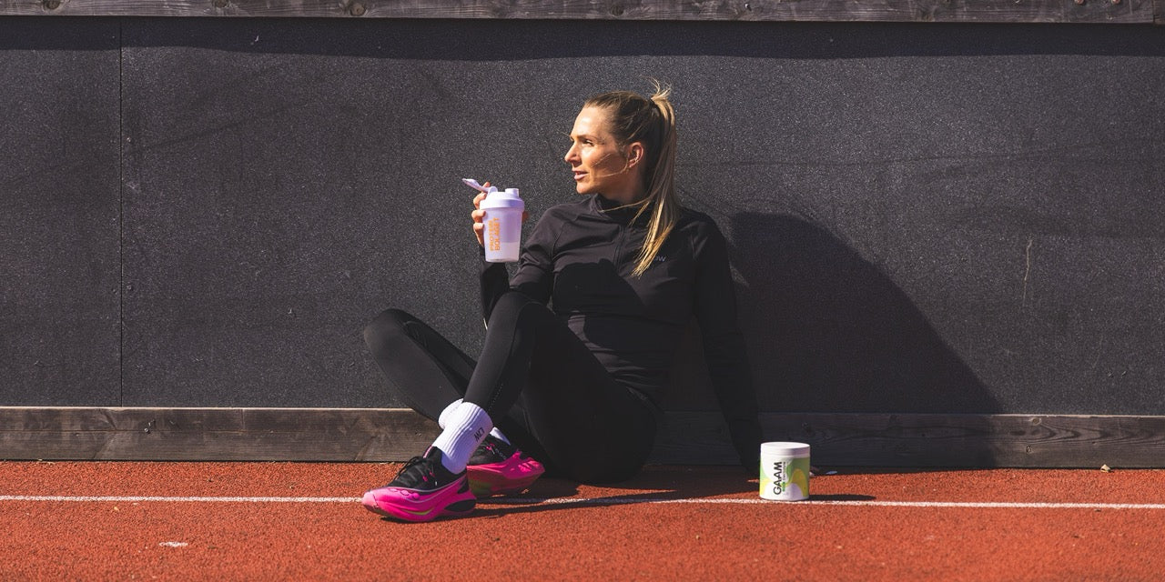 Kvinna i löparträning sitter på friidrottsbana med proteinshaker i handen och GAAM kosttillskott bredvid sig